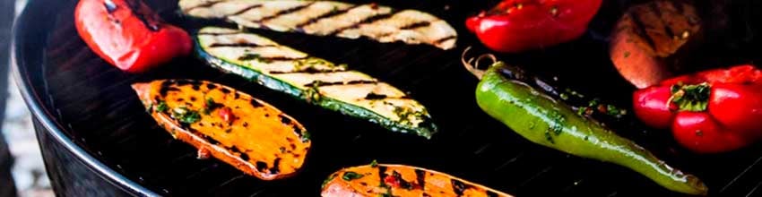Rød paprika, gresskar, squash og en halv løk tilberedes på en liten, rund grill på et trebord.