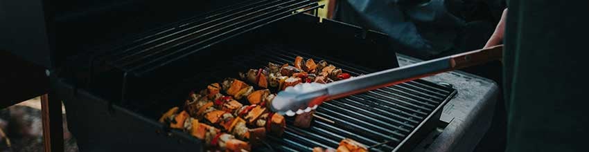 Grillspyd tilberedes på gassgrill på terrassen i dempet kveldssol, med person i gang med matlaging.