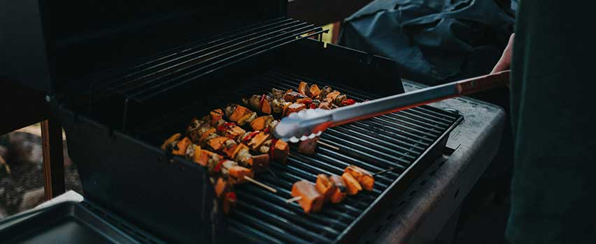Syv grillspyd med grønnsaker vendes med grilltang på en stor, firkantet, varm grill.