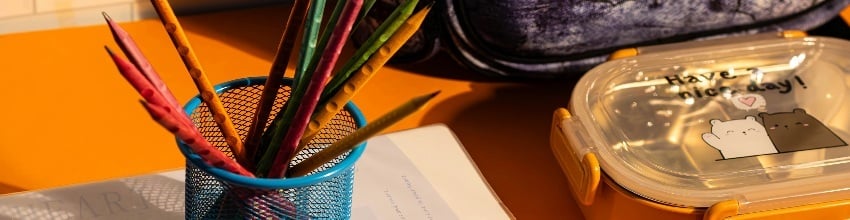 Kopp med fargeblyanter, notatbok og matboks på et oransje skrivebord med en skolesekk i bakgrunnen.
