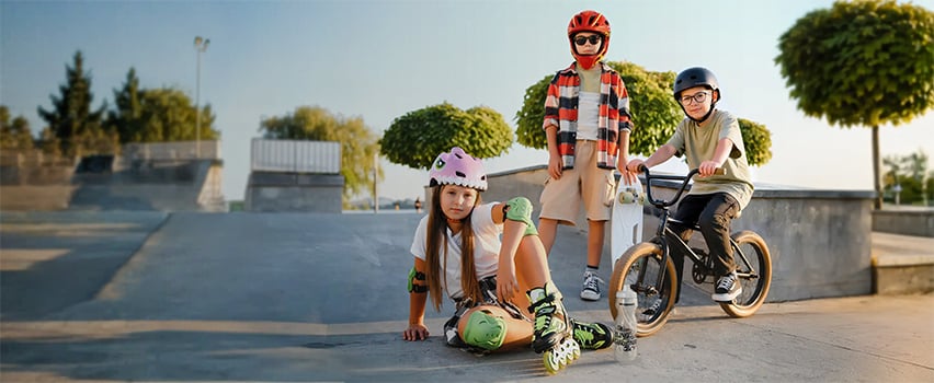 Tre barn med hjelmer og verneutstyr fra Wozinsky på skateboard, rulleskøyter og BMX-sykkel i skatepark