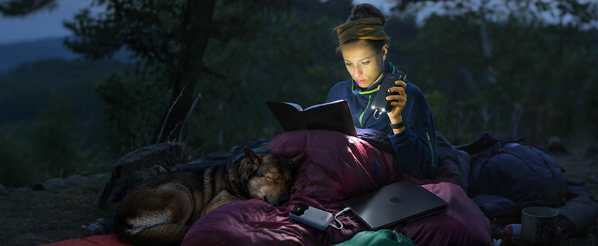 Kvinne leser bok i telt med hund og lader smarttelefon og laptop med Wozinsky powerbank og pannelykten
